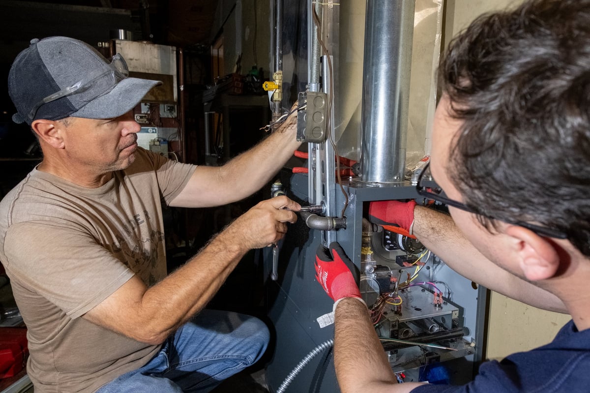 Two technicians work on installing or repairing a heating system component in a residential HVAC unit
