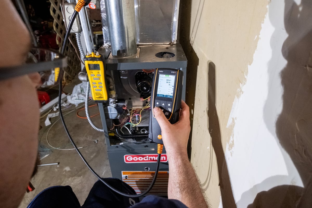 Technician holding digital meter while servicing HVAC unit with exposed wiring and components