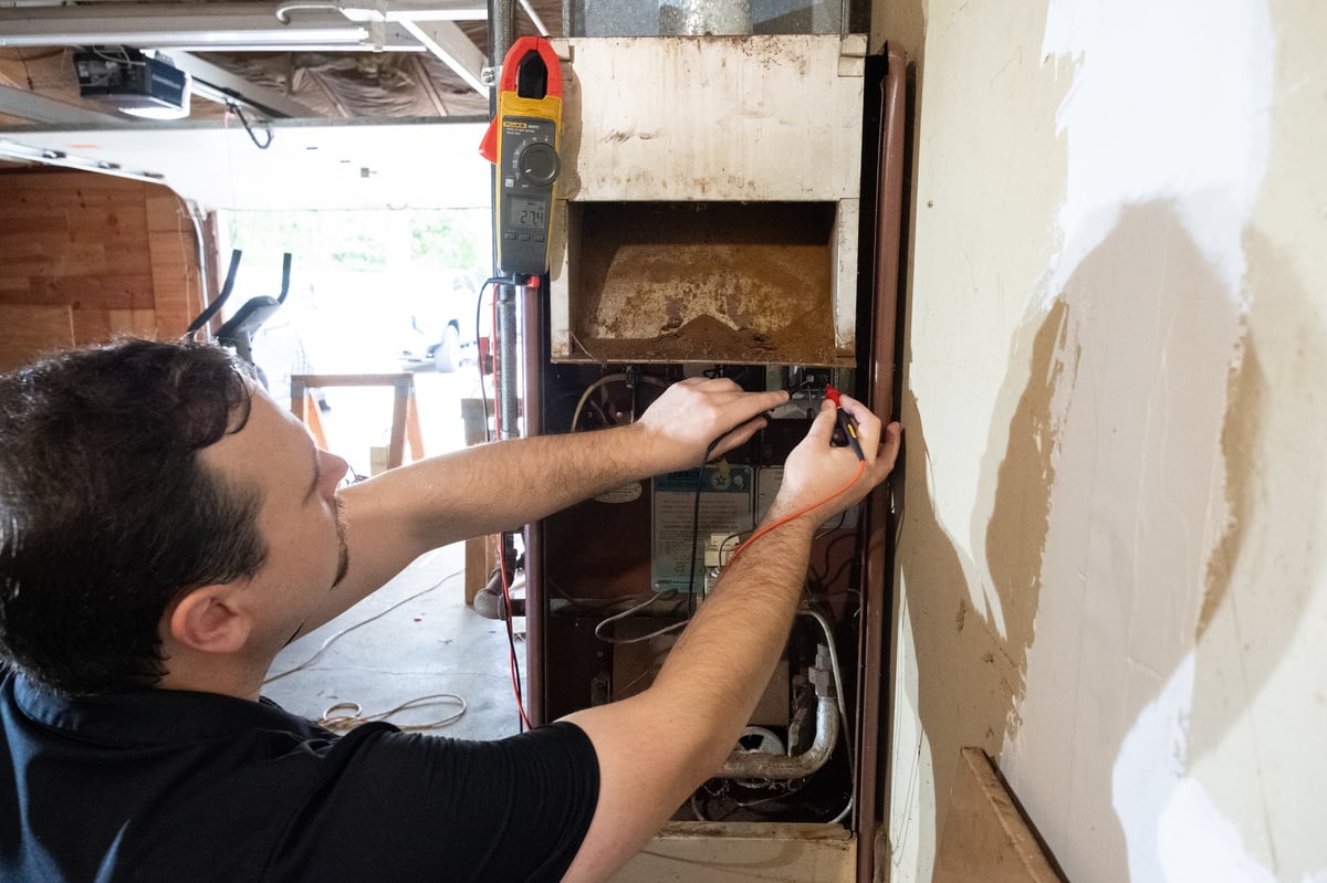 Technician using a multimeter to test an electrical panel mounted on a wall during maintenance work